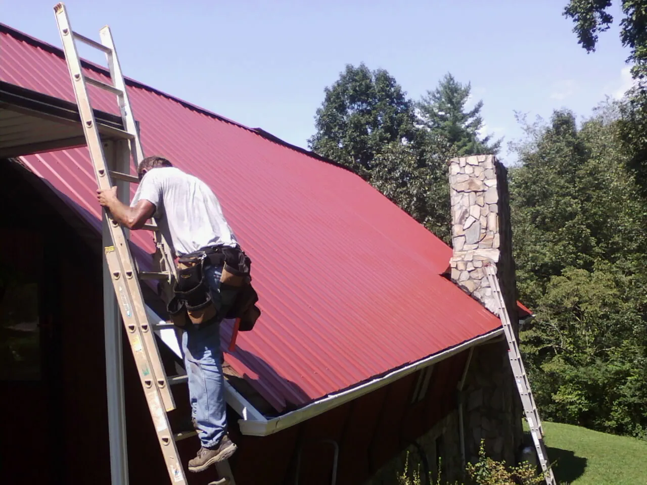 Roofer on a ladder beside a red metal roof and stone chimney — Soffit & Fascia Repair by Roofing Brothers in Andrews Air Force Base
